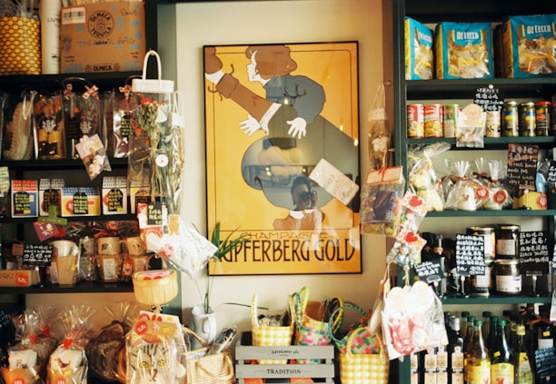 A small store interior filled with various packaged goods displayed on dark shelves. A prominent vintage poster with the words 'Champagne Kupferberg Gold' is centered on the wall. The shelves are stocked with snacks, pasta, condiments, and decorative items, many labeled with tags. Brightly colored baskets and gift bags are arranged at the bottom.