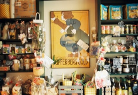 A small store interior filled with various packaged goods displayed on dark shelves. A prominent vintage poster with the words 'Champagne Kupferberg Gold' is centered on the wall. The shelves are stocked with snacks, pasta, condiments, and decorative items, many labeled with tags. Brightly colored baskets and gift bags are arranged at the bottom.