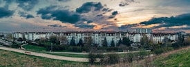 A panoramic view of a residential area with several mid-rise apartment buildings. The sky is partly cloudy with a colorful sunset, casting a warm glow over the scene. In the foreground, a grassy area and pathway are visible, lined with benches and sparse trees.