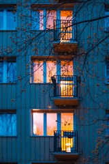 An exterior view of a residential building with illuminated windows glowing warmly in contrast to the bluish tint of the surrounding wall. Several balconies are visible, partially obscured by leafless tree branches in the foreground.