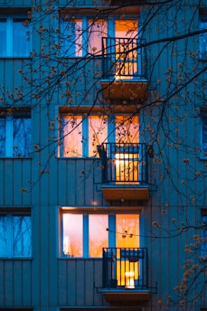 An exterior view of a residential building with illuminated windows glowing warmly in contrast to the bluish tint of the surrounding wall. Several balconies are visible, partially obscured by leafless tree branches in the foreground.