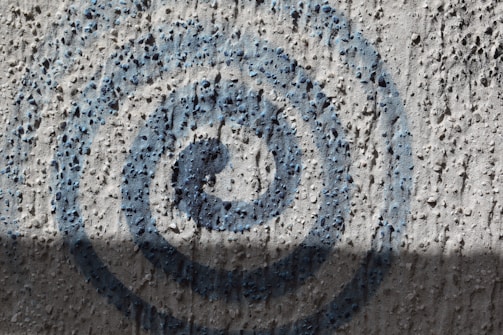 A textured concrete wall with a spiral pattern spray-painted in blue. The surface has small protrusions and shadows that add depth to the image.