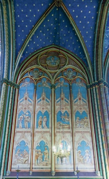 A richly detailed stained glass panel featuring religious imagery set within pointed arches. The artwork is framed by ornate architectural designs, and a chandelier is visible in the foreground. The blue color dominates the background, contrasted by intricate patterns and figures.