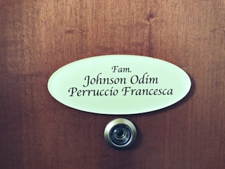 A wooden door features an oval nameplate with black text displaying names, and a metal doorbell or peephole is situated below it.