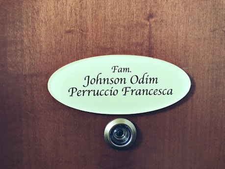 A wooden door features an oval nameplate with black text displaying names, and a metal doorbell or peephole is situated below it.