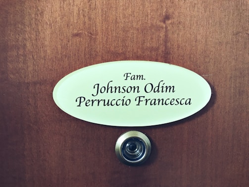 A wooden door features an oval nameplate with black text displaying names, and a metal doorbell or peephole is situated below it.
