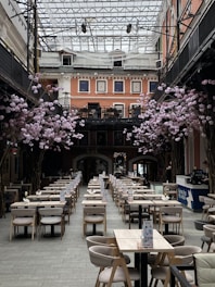 A spacious indoor dining area with rows of neatly arranged tables and chairs. Pink floral arrangements decorate the space, and the high glass ceiling allows natural light to illuminate the room. The surrounding architecture features multiple stories with salmon-colored walls and large windows.