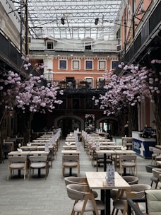 A spacious indoor dining area with rows of neatly arranged tables and chairs. Pink floral arrangements decorate the space, and the high glass ceiling allows natural light to illuminate the room. The surrounding architecture features multiple stories with salmon-colored walls and large windows.