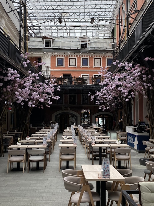 A spacious indoor dining area with rows of neatly arranged tables and chairs. Pink floral arrangements decorate the space, and the high glass ceiling allows natural light to illuminate the room. The surrounding architecture features multiple stories with salmon-colored walls and large windows.