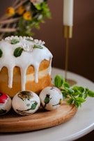 A baked cake topped with white icing and decorative green leaves sits on a wooden board. Three painted eggs with floral designs are placed beside the cake. A lit white candle in a gold holder and some fresh green leaves are visible in the background, adding a festive and cozy atmosphere.