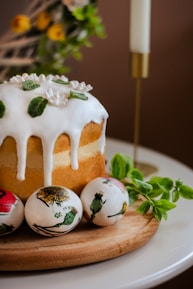 A baked cake topped with white icing and decorative green leaves sits on a wooden board. Three painted eggs with floral designs are placed beside the cake. A lit white candle in a gold holder and some fresh green leaves are visible in the background, adding a festive and cozy atmosphere.