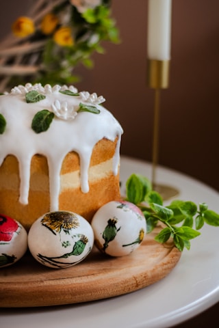 A baked cake topped with white icing and decorative green leaves sits on a wooden board. Three painted eggs with floral designs are placed beside the cake. A lit white candle in a gold holder and some fresh green leaves are visible in the background, adding a festive and cozy atmosphere.