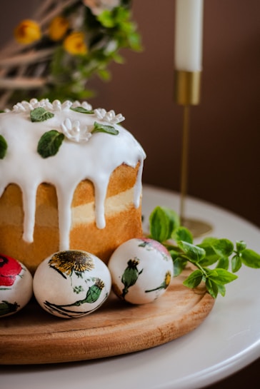 A baked cake topped with white icing and decorative green leaves sits on a wooden board. Three painted eggs with floral designs are placed beside the cake. A lit white candle in a gold holder and some fresh green leaves are visible in the background, adding a festive and cozy atmosphere.
