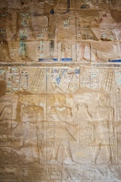 Ancient Egyptian hieroglyphs and relief carvings on a stone wall featuring figures with headdresses, symbols, and detailed attire. The artwork includes registers filled with various intricate symbols in subtle hues.