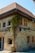 A rustic building with a stone facade and a red tiled roof, adorned with vintage-style lamps. A climbing plant with yellow flowers grows along the wall from a large ceramic pot. The windows and doors have wooden frames, and there are subtle textures on the walls.