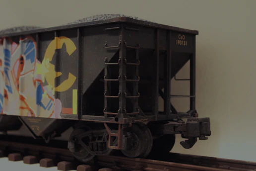 A freight train car on railway tracks, featuring colorful graffiti art with abstract designs and bright colors. The train car has a dark, weathered appearance, conveying a sense of urban grit.