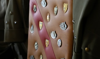 A collection of various badges and insignias is displayed on a brown leather sash or strap. The badges include a mix of shield shapes and circular designs, each with intricate details such as emblems, coats of arms, and figures. The background contains army green-colored fabric, possibly part of a military uniform, adding context to the badges.