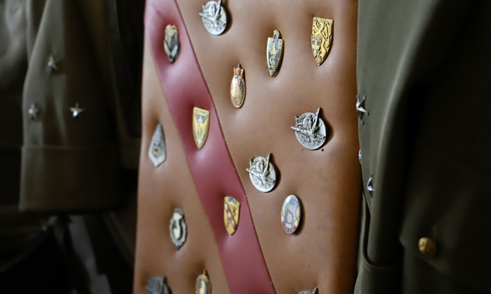 A collection of various badges and insignias is displayed on a brown leather sash or strap. The badges include a mix of shield shapes and circular designs, each with intricate details such as emblems, coats of arms, and figures. The background contains army green-colored fabric, possibly part of a military uniform, adding context to the badges.