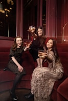 Three women are seated on a red velvet couch, each holding a glass with a golden drink, wearing elegant attire. One woman is wearing a dark floral dress, another is adorned in a black outfit, and the third is in a patterned dress with sequins. They are posing with cheerful expressions in a warmly lit, luxurious setting.