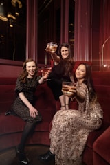 Three women are seated on a red velvet couch, each holding a glass with a golden drink, wearing elegant attire. One woman is wearing a dark floral dress, another is adorned in a black outfit, and the third is in a patterned dress with sequins. They are posing with cheerful expressions in a warmly lit, luxurious setting.