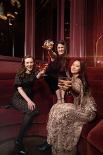 Three women are seated on a red velvet couch, each holding a glass with a golden drink, wearing elegant attire. One woman is wearing a dark floral dress, another is adorned in a black outfit, and the third is in a patterned dress with sequins. They are posing with cheerful expressions in a warmly lit, luxurious setting.