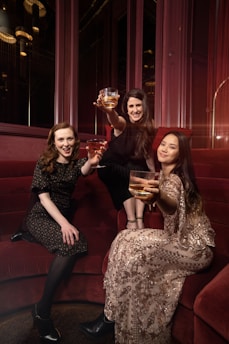 Three women are seated on a red velvet couch, each holding a glass with a golden drink, wearing elegant attire. One woman is wearing a dark floral dress, another is adorned in a black outfit, and the third is in a patterned dress with sequins. They are posing with cheerful expressions in a warmly lit, luxurious setting.