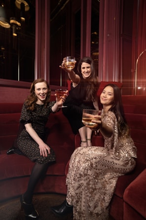 Three women are seated on a red velvet couch, each holding a glass with a golden drink, wearing elegant attire. One woman is wearing a dark floral dress, another is adorned in a black outfit, and the third is in a patterned dress with sequins. They are posing with cheerful expressions in a warmly lit, luxurious setting.