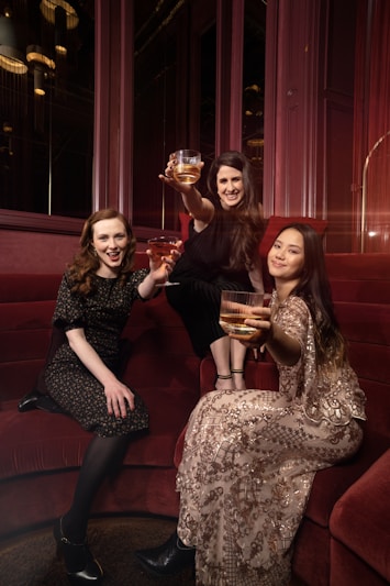 Three women are seated on a red velvet couch, each holding a glass with a golden drink, wearing elegant attire. One woman is wearing a dark floral dress, another is adorned in a black outfit, and the third is in a patterned dress with sequins. They are posing with cheerful expressions in a warmly lit, luxurious setting.