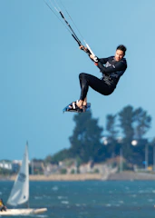 A man is engaged in kiteboarding, soaring high in the air with a kite. He is wearing a wetsuit and appears to be skillfully controlling the kite. The background includes a large body of water with a sailboat, and distant trees and buildings under a clear blue sky.