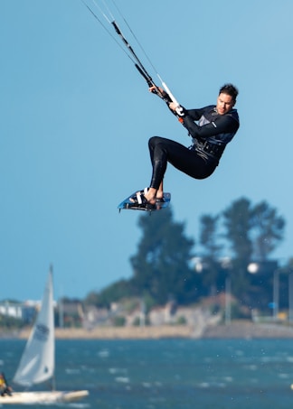 A man is engaged in kiteboarding, soaring high in the air with a kite. He is wearing a wetsuit and appears to be skillfully controlling the kite. The background includes a large body of water with a sailboat, and distant trees and buildings under a clear blue sky.