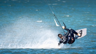 A person is engaged in kite surfing on a body of water, performing a dynamic maneuver with water splashing around them. They are wearing a wetsuit and are attached to a kite that is pulling them through the air.