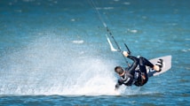 A person is engaged in kite surfing on a body of water, performing a dynamic maneuver with water splashing around them. They are wearing a wetsuit and are attached to a kite that is pulling them through the air.