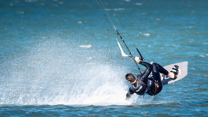 A person is engaged in kite surfing on a body of water, performing a dynamic maneuver with water splashing around them. They are wearing a wetsuit and are attached to a kite that is pulling them through the air.
