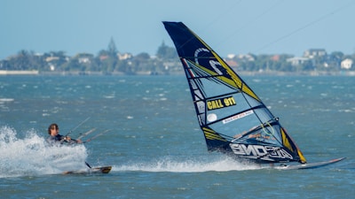 A person is engaged in kite surfing across a body of water, with a colorful sail that has text and patterns on it. The water is splashing around the surfer as they navigate. The background shows a distant shoreline with trees and buildings under a clear blue sky.