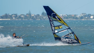 A person is engaged in kite surfing across a body of water, with a colorful sail that has text and patterns on it. The water is splashing around the surfer as they navigate. The background shows a distant shoreline with trees and buildings under a clear blue sky.