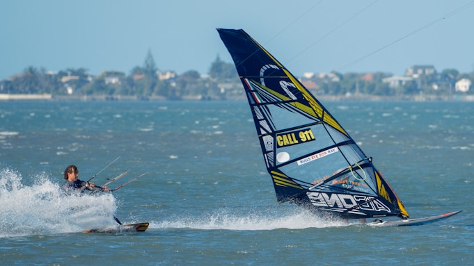 A person is engaged in kite surfing across a body of water, with a colorful sail that has text and patterns on it. The water is splashing around the surfer as they navigate. The background shows a distant shoreline with trees and buildings under a clear blue sky.