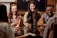 A group of four friends are sitting around a polished wooden table, laughing and enjoying drinks in a cozy, stylish setting. The group appears to be engaged in a warm and lively conversation, creating an atmosphere of camaraderie and joy.