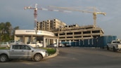 A construction site with a partially built multi-story building and two large cranes. Several vehicles, including cars and trucks, are parked along a pavement and in front of a building labeled 'Ashton Tower Property Group'. The sky is cloudy with faint sunlight casting shadows.