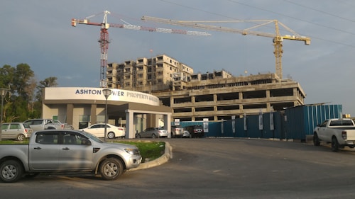 A construction site with a partially built multi-story building and two large cranes. Several vehicles, including cars and trucks, are parked along a pavement and in front of a building labeled 'Ashton Tower Property Group'. The sky is cloudy with faint sunlight casting shadows.