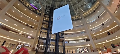 A spacious and modern shopping mall interior featuring multiple floors with numerous stores. Prominent elements include a large vertical digital screen in the center and escalators on either side. Bright lighting and sleek architectural design with a high atrium contribute to the open atmosphere.