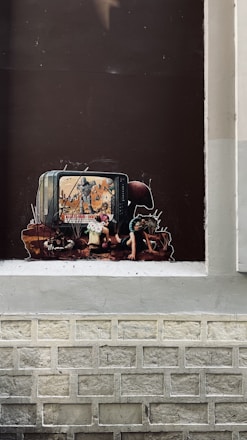 A piece of street art depicts a television with a scene of soldiers and a destroyed landscape on the screen. In front of it, a child's figure is positioned, appearing absorbed by the screen's contents. The surrounding imagery includes dark and muted colors, giving a grim and thought-provoking atmosphere.