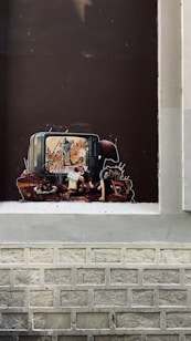 A piece of street art depicts a television with a scene of soldiers and a destroyed landscape on the screen. In front of it, a child's figure is positioned, appearing absorbed by the screen's contents. The surrounding imagery includes dark and muted colors, giving a grim and thought-provoking atmosphere.