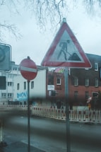 A street scene featuring several road signs including a no entry sign and a construction warning sign. In the background, there are buildings with graffiti on the walls, and a construction area with barricades. Bare tree branches can be seen above.