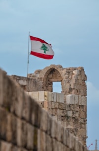 An old stone structure with an archway is topped by a flagpole flying the Lebanese flag against a clear blue sky.