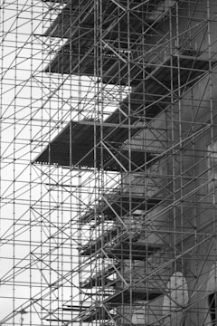 A large, complex scaffold structure is constructed against a building. The metal framework of the scaffolding is prominent, creating a grid-like pattern with horizontal and vertical lines. Several platforms are visible within the scaffolding, indicating areas for workers to stand. The scene conveys a sense of industrial organization and activity.