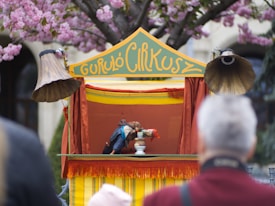 A small puppet theater setup outdoors features a colorful stage with yellow and red drapes. Two vintage-style gramophone horns flank the stage. The sign above reads 'Guruló Cirkusz'. Puppets are actively engaged in performance inside the theater. The scene is surrounded by blooming cherry blossom trees, with an audience watching in the foreground.