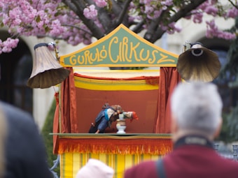 A small puppet theater setup outdoors features a colorful stage with yellow and red drapes. Two vintage-style gramophone horns flank the stage. The sign above reads 'Guruló Cirkusz'. Puppets are actively engaged in performance inside the theater. The scene is surrounded by blooming cherry blossom trees, with an audience watching in the foreground.