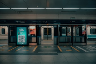 An empty subway platform features illuminated advertising, with a QR code visible in the center. Subway doors are open, revealing the interior seating area. The signage above the platform indicates directions and locations in a transport system.