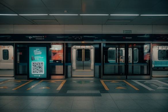 An empty subway platform features illuminated advertising, with a QR code visible in the center. Subway doors are open, revealing the interior seating area. The signage above the platform indicates directions and locations in a transport system.