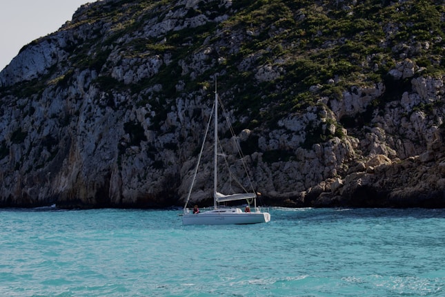 A sailboat is floating on vibrant turquoise water near a large, rocky cliff covered in patches of greenery. The sunlight highlights the rugged texture of the rocks and the calmness of the water under a clear sky.